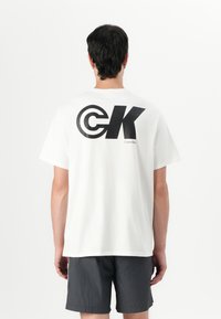 Balta medvilninė marškinėliai su dideliu juodu "CK" logotipu ant nugaros ir "Calvin Klein" tekstu, derinami su tamsiai pilkais šortais. Paprastas, laisvas kirpimas.