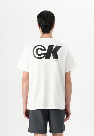White cotton t-shirt with a large black "CK" logo on the back and "Calvin Klein" text, paired with dark grey shorts. Simple, relaxed fit.
