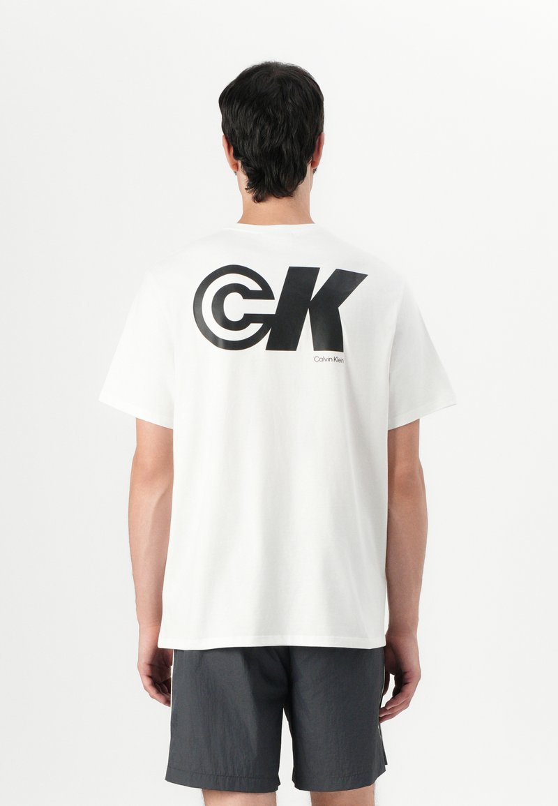 Balta medvilninė marškinėliai su dideliu juodu "CK" logotipu ant nugaros ir "Calvin Klein" tekstu, derinami su tamsiai pilkais šortais. Paprastas, laisvas kirpimas.