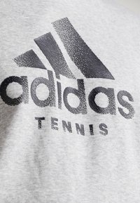 Camiseta gris con un prominente logo negro de Adidas con detalles texturizados. "TENNIS" impreso debajo en una fuente estilizada y punteada.