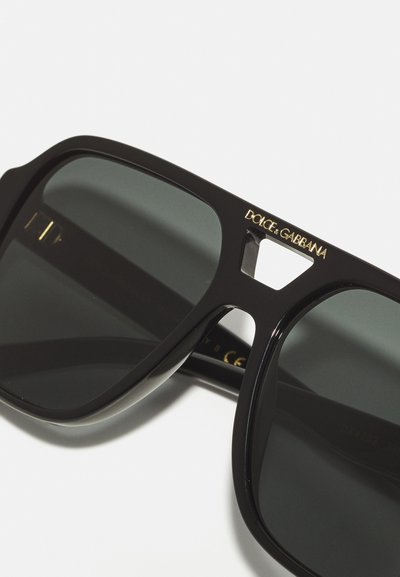 Juodi akiniai nuo saulės su storu rėmeliu, turintys pilkus lęšius ir auksinį „DOLCE & GABBANA“ logotipą ant šventyklų. Kampuotas dizainas su išpjovų detale.