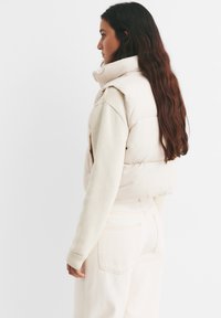 Svijetlo bež kviltani prsluk s visokim ovratnikom, u kombinaciji s kremasta puloverom i svijetlo bež trapericama visokog struka. Glatka tekstura, opušteni kroj.