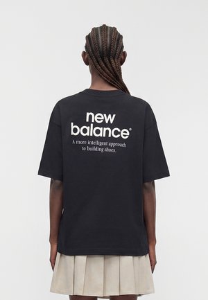 Άτομο με πλεξούδες, που φοράει μαύρο T-shirt New Balance με λευκό κείμενο και μπεζ φούστα με πιέτες, γυρισμένο με την πλάτη στην κάμερα.