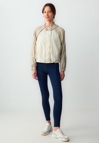 Leichte beige Kapuzenjacke mit elastischen Bündchen, kombiniert mit dunkelblauen Leggings und weißen Sneakers mit grauen Akzenten.
