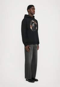 Joven de pie vestido con una sudadera gráfica negra, pantalones vaqueros anchos y desvaídos en negro, y zapatos de cuero negros, frente a un fondo simple.