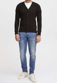 Mężczyzna w czarnej zapinanej kardiganie, białej koszulce pod, niebieskich dżinsach i białych sneakersach stoi na tle plain white.