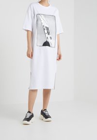 Biała sukienka t-shirtowa z graficznym nadrukiem z napisem "DKNY" i gwiazdkami. Krótkie rękawy, boczne rozporki, w zestawieniu z czarnymi butami sportowymi. Gładka tekstura materiału.