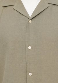 Ελαφρύ πράσινο πουλόβερ ελιάς με γιακά πέταλο και τέσσερα μπεζ κουμπιά κλειστά πάνω από ένα λευκό υποκάμισο.