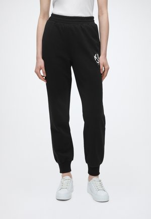 Personne portant un pantalon de jogging noir avec des poignets élastiques et des baskets blanches sur un fond clair uni.