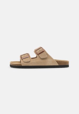 Tan suede slide sandal with two adjustable straps, featuring metallic buckles, a cork footbed, and a textured black rubber sole.
