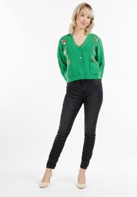 Cardigan verde a maglia con scollo a V, bottoni frontali e due tasche; presenta ricami floreali dorati sul lato sinistro. Jeans skinny neri.