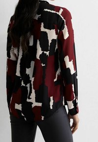 Blusa a maniche lunghe in nero, bordeaux e crema. Presenta un motivo astratto, tessuto morbido e un dettaglio con bottone sul polsino.