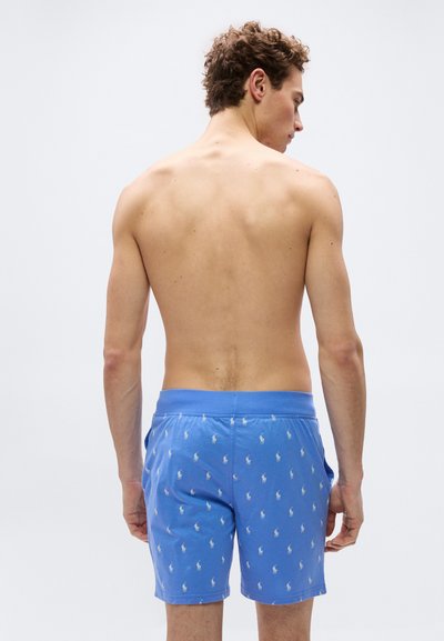 Άνδρας με σγουρά μαλλιά, φανερός από πίσω, φοράει μπλε μαγιό σορτς με λευκό επαναλαμβανόμενο μοτίβο αλόγου και αναβάτη σε απλό φόντο.