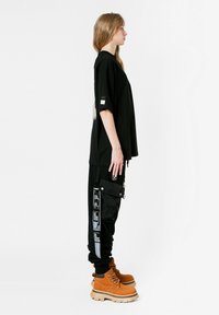 Camiseta negra de gran tamaño con diseño gráfico en la parte posterior, combinada con pantalones cargo negros con bolsillos laterales y detalles en texto blanco. Botas beige.