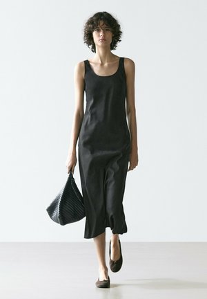 Donna che cammina indossando un vestito midi nero senza maniche, tiene in mano una borsa nera testurizzata e indossa scarpe basse scure, su uno sfondo chiaro e uniforme.