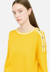 Maglione giallo con strisce bianche nel colletto e spalle, caratterizzato da bottoni decorativi dorati. Texture morbida, vestibilità rilassata, design casual.