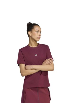 Une femme portant un tee-shirt Adidas bordeaux et un short assorti se tient debout, les bras croisés, sur un fond blanc uni.