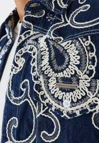Giacca di denim con motivi paisley ricamati bianchi, chiusura a bottoni e etichetta con marchio. Tessuto blu scuro con texture di cucitura visibile.