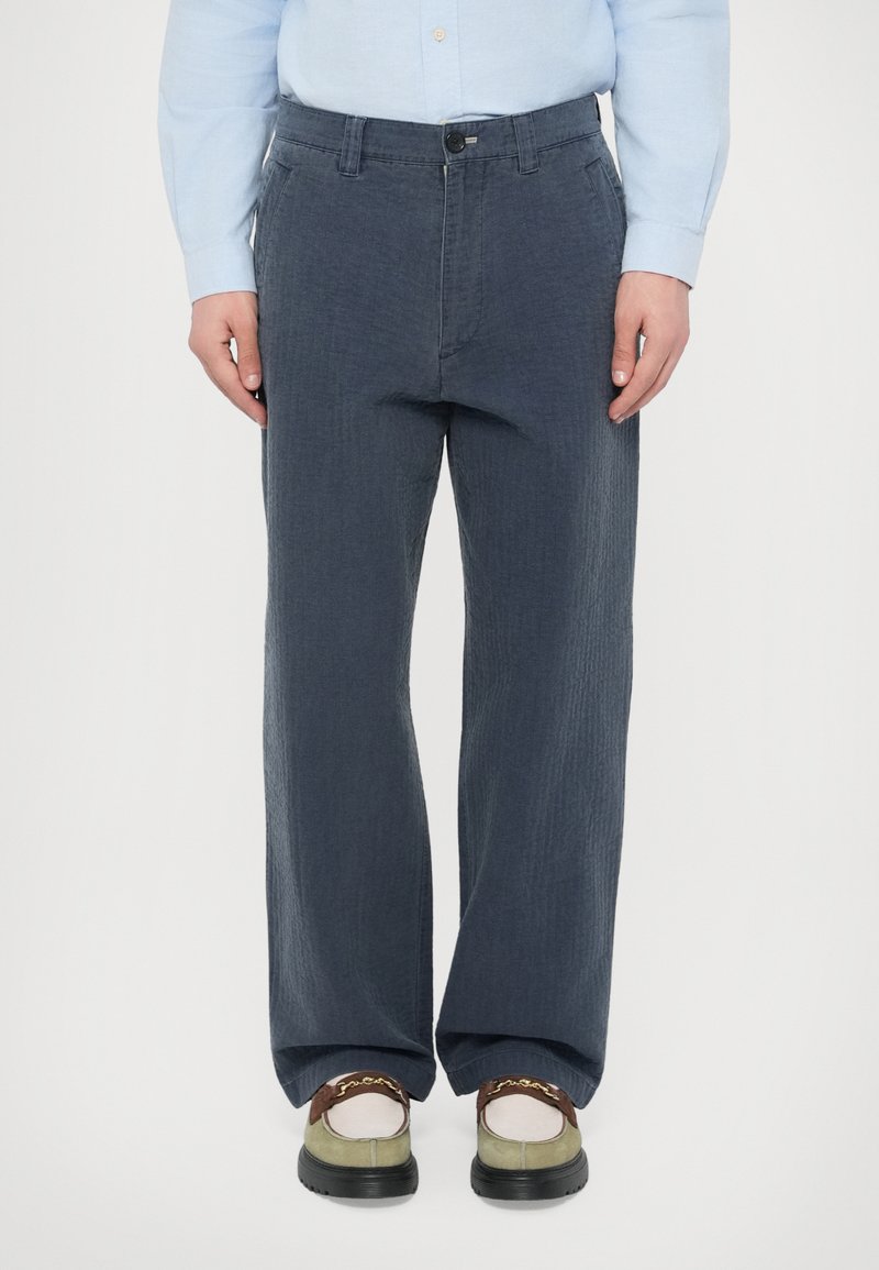 Ελαφριά, σκούρα γκρι παντελόνια με φαρδιά μπατζά�κια και υφή, με πλαϊνές τσέπες και κλείσιμο με κουμπιά, φορεμένα με casual παπούτσια.