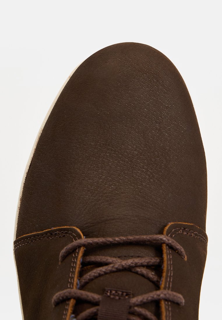 Zapato de cuero marrón con una textura suave, puntera redondeada y costuras en contraste color beige. Presenta cordones marrones y una suela de goma ligera.