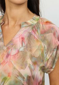 Genomskinlig blus med blommönster i rosa, gröna och beige nyanser, med V-ringning och mjukt, flödande tyg. Korta ärmar och lätt lyster.