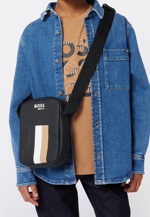 Borsa a tracolla nera con strisce bianche e beige, realizzata in materiale resistente. Caratterizzata da un logo e da una tracolla regolabile. Indossata con una camicia di jeans e una t-shirt marrone.