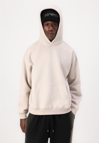 Lys beige hættetrøje med en frontlomme, justerbar hætte og ribbede cuffs. Bærers med sorte joggers og en sort beanie med bogstaver.