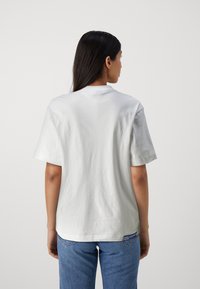 Camiseta de algodón blanca con mangas cortas, escote redondo y una textura suave, mostrada desde la parte de atrás, combinada con jeans de mezclilla azules.