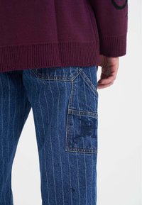 Fioletowy dzianinowy sweter z prążkowanym dołem, w połączeniu z niebieskimi spodniami dżinsowymi w paski. Spodnie mają dużą kieszeń naszywaną z haftowanym logo.