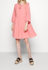 Rosa Leinenkleid mit gestuftem Design, weiten Ärmeln und einer Bindung am Hals. Kombiniert mit schwarzen Plateau-Sandalen. Einfacher, strukturierter Stoff.