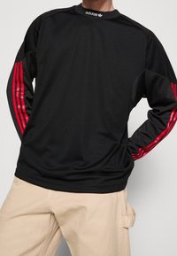 Czarna koszulka Adidas z długim rękawem i czerwonymi paskami na rękawach, noszona z beżowymi spodniami, ręce w kieszeniach koszulki.