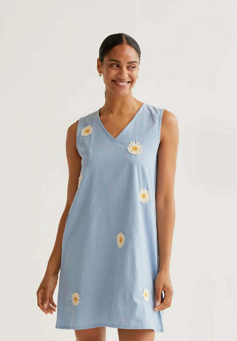 Kvinde med mørkt hår iført en lyseblå, ærmeløs kjole med hvid margueritemotiv, smilende og stående mod en ensfarvet lys baggrund.