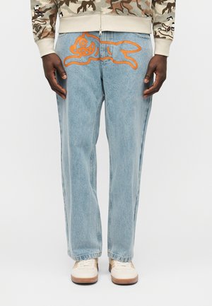 Lyseblå jeans med en orange grafik af en løbende tegneseriehund over de øverste forreste lår, båret med hvide sneakers og camouflagejakke.