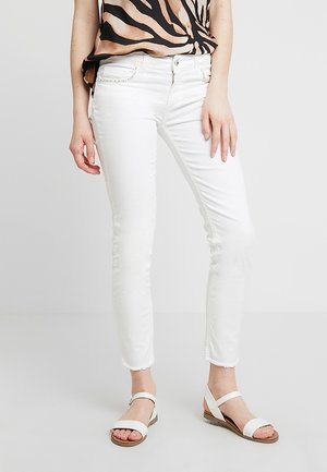 Frau trägt weiße, schlanke Jeans mit ausgefransten Säumen, weiße Sandalen mit Knöchelriemen und ein Zebra-Print-Oberteil, steht vor einem schlichten Hintergrund.