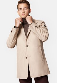 Beżowy trencz z wyraźnym kołnierzem, trzema guzikami i bocznymi kieszeniami, w połączeniu z brązowym golfem o prążkowanej teksturze. Gładka faktura.