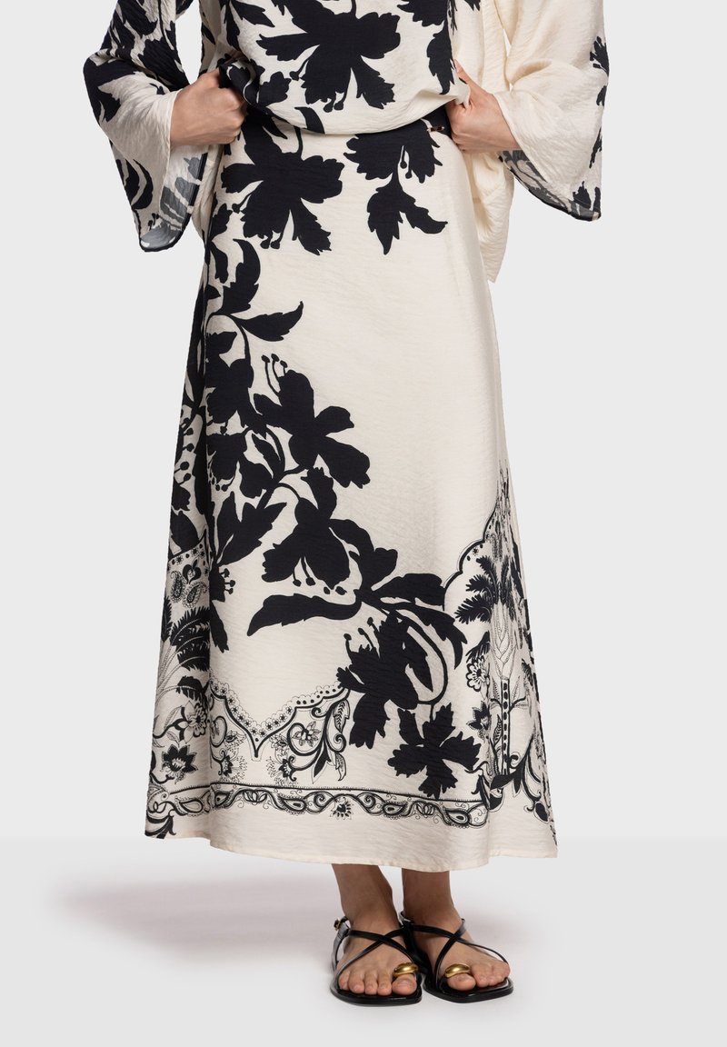 Falda hasta la rodilla con estampado floral negro sobre fondo beige, con detalles delicados en los bordes y textura suave. Combinada con sandalias planas.