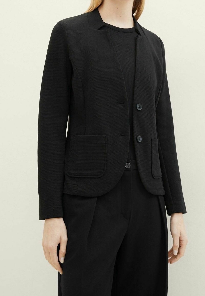 Femme portant un blazer noir ajusté avec deux boutons et des poches plaquées, associé à un pantalon noir et un top noir.