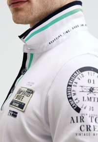 Weißes Poloshirt mit schwarz und türkis gestreiftem Kragen, das mit Patches und gedrucktem Text versehen ist. Der Text in der Nähe des Kragens lautet "BOARDING TIME: 0900 AM."