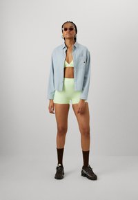 Chemise en jean bleu clair avec un tissu texturé, accompagnée d'un soutien-gorge de sport vert fluo et de shorts, complétée par des baskets sombres et épaisses et des chaussettes marron.