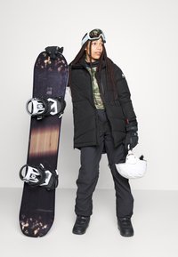 Snowboarder in een zwarte pufferjack, camouflage top, zwarte broek en handschoenen. Houdt een snowboard vast met witte bindingen en een witte helm.