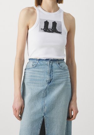 Femme portant un haut blanc sans manches avec un motif de bottes de cowboy noires et une jupe en denim bleu clair taille haute avec une fente à l'avant.