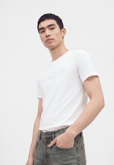 Jeune homme portant un t-shirt blanc à manches courtes et un jean gris, une main dans la poche, debout devant un fond uni.