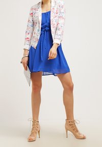 Femme portant une robe bleue avec un blazer floral, tenant une tablette dans une main et portant des chaussures à lacets beiges à talons hauts.