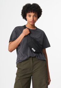 Schwarze Stoffgürteltasche mit einer vorderen Reißverschlusstasche und einem Logotag. Das Model trägt ein graues, übergroßes T-Shirt und grüne Hosen.