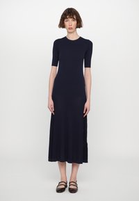 Abito in maglia a coste blu navy con maniche corte e corpetto aderente, che scende fino a una lunghezza midi, abbinato a scarpe basse in pelle marrone scuro.