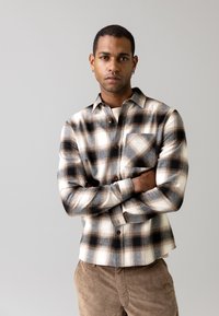 Camicia a quadri nei toni beige e neri, realizzata in tessuto morbido, con chiusura frontale a bottoni, colletto e taschino sul petto. Aspetto texturizzato.