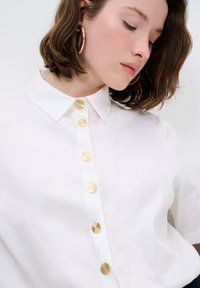 Camicia bianca a maniche corte con colletto a punta e sei bottoni metallici dorati sul davanti, caratterizzata da un tessuto liscio e strutturato.
