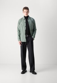 Camisa verde claro de botones sobre un cuello de tortuga negro, combinada con pantalones oscuros y zapatos negros. Tela suave y diseño de corte recto.