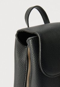 Μαύρο δερμάτινο σακίδιο με υφή επιφάνειας, καμπυλωτό καπάκι και λεπτομέρεια χρυσλού φερμουάρ. Διαθέτει επάνω χειρολαβή για εύκολη μεταφορά.