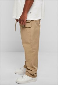 Beige cargo pants with side pockets, made of cotton blend fabric, featuring a relaxed fit and adjustable drawstring at the waist. White sneakers.